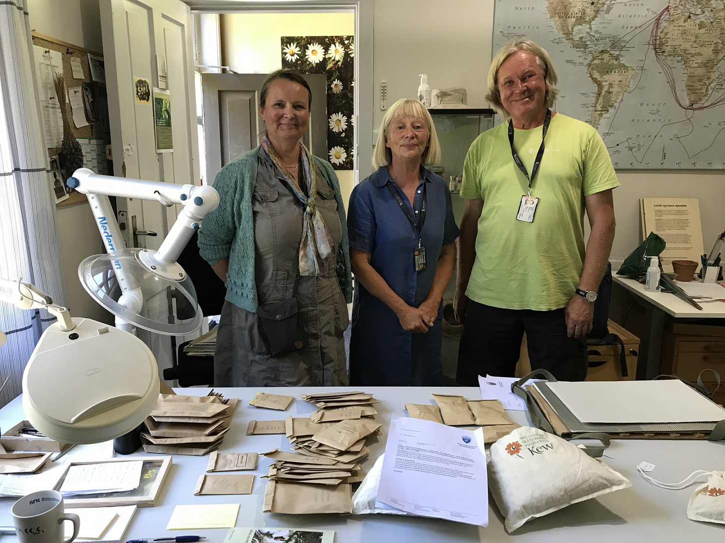 3 people standing in a row smiling behind a work bench. The work bench has a variety of paper envelopes and cloth bags on it along with a stack of herbarium specimens